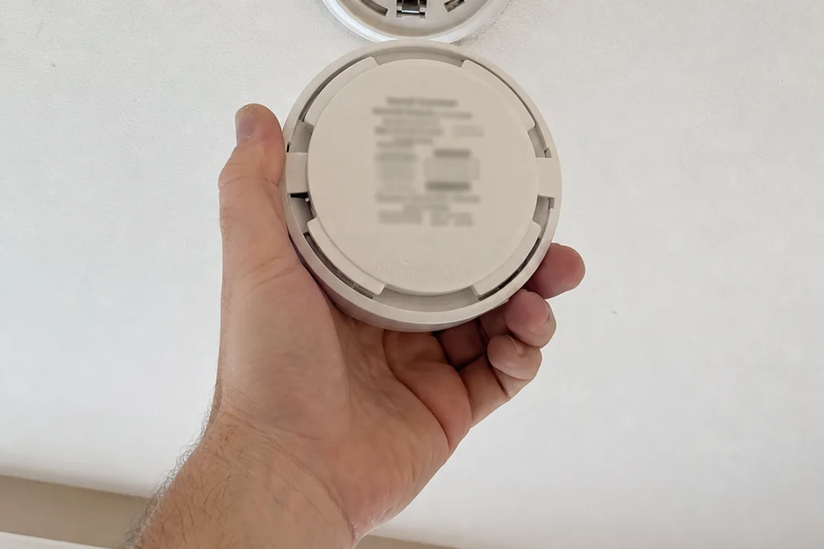 Close view of a bare hand twisting a round smoke alarm free of its ceiling bracket and turning it over, a printed date label showing as a soft blurred impression on the back, the empty bracket visible above in even daylight