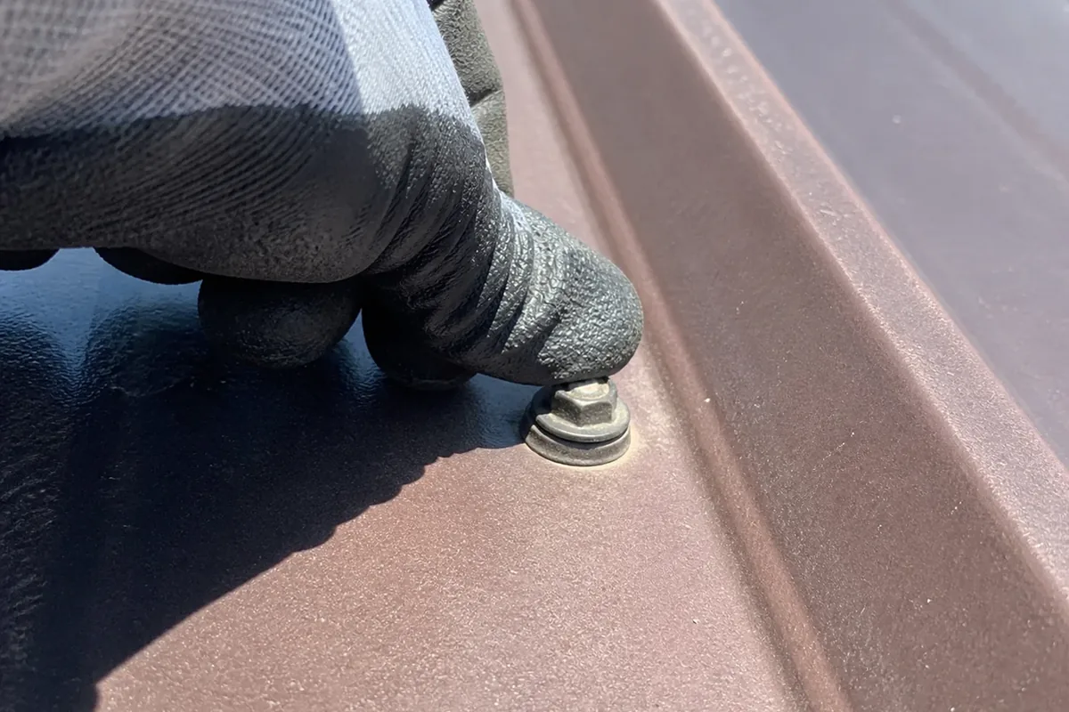 A hand pressing a gloved thumb against a backed-out roofing screw head to test for movement on an exposed-fastener panel