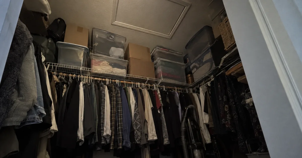 A bedroom closet with an attic access hatch in the ceiling, the wire shelf beneath it stacked to the ceiling with plastic bins and cardboard boxes above a packed clothes rod