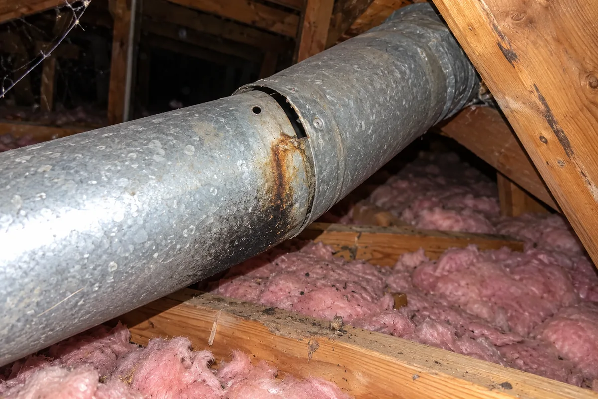 A galvanized flue pipe in an attic with a separated joint, a rust streak and soot stain beside the gap, and pink insulation below
