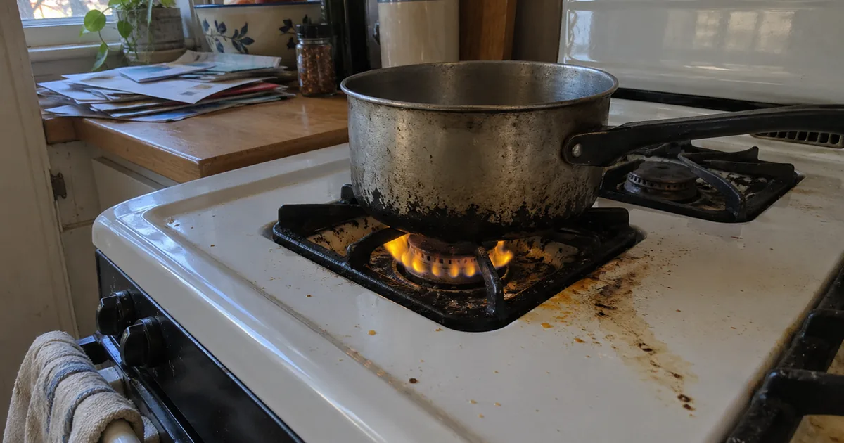 A gas range burner with a lazy yellow-orange flame under a soot-darkened saucepan on a grease-stained cooktop in dim window light