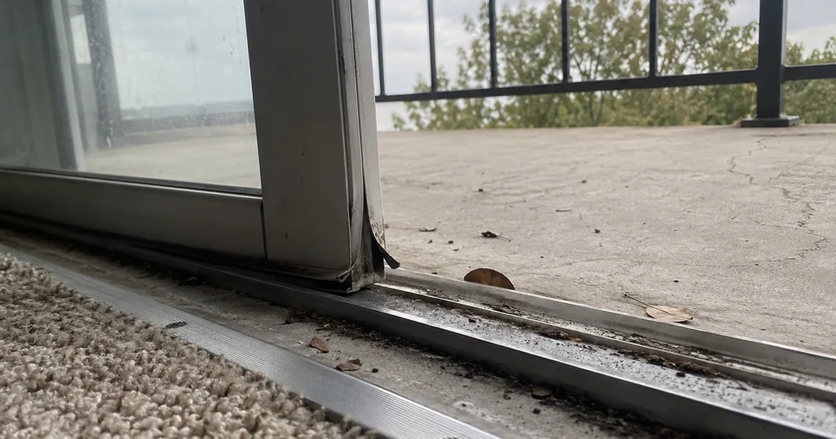 Low view of a condo sliding patio door open a few inches, with grit and leaves in the aluminum track, a split weatherstrip at the door corner and a cracked concrete balcony beyond