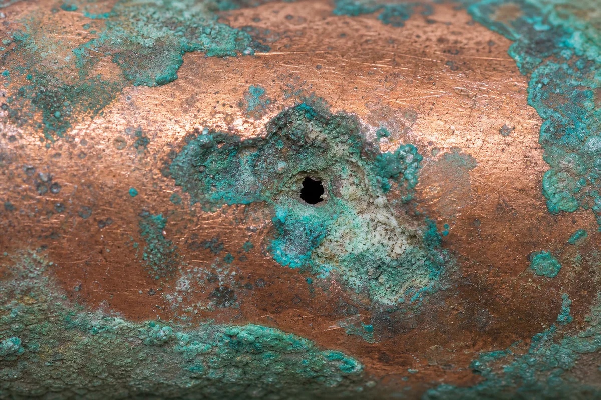 Extreme macro of a copper pipe surface showing green and turquoise pitting corrosion, small craters and a tiny dark pinhole eaten through the copper wall