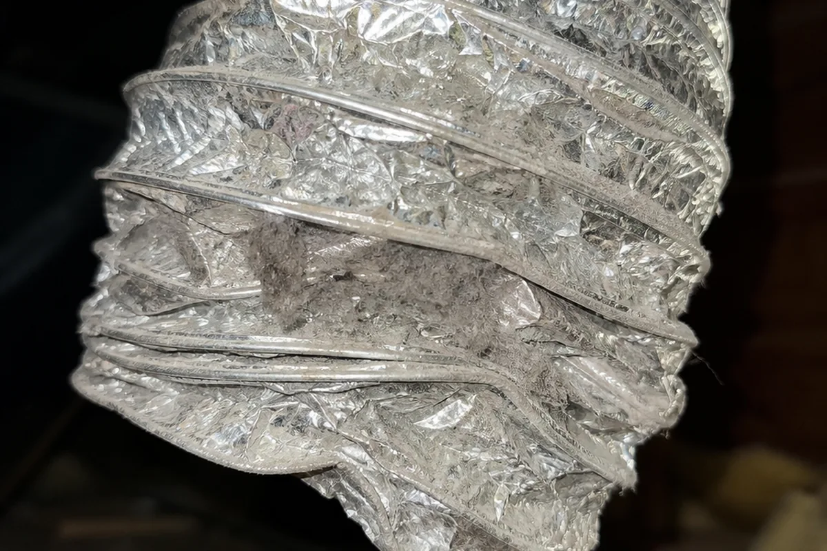 A crushed foil flex dryer duct behind a dryer pulled away from the wall, the duct visibly flattened where it was compressed against framing