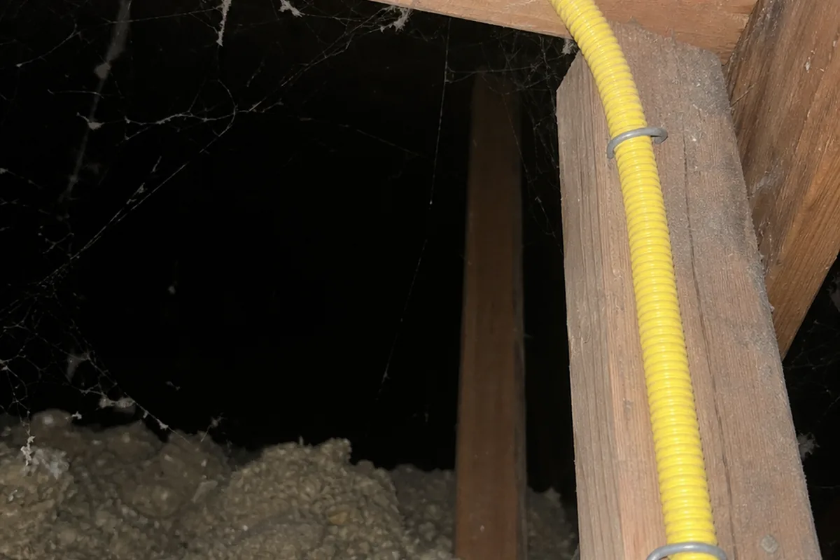 Yellow CSST gas tubing threaded through drilled holes in dusty attic roof framing under hard on-camera flash