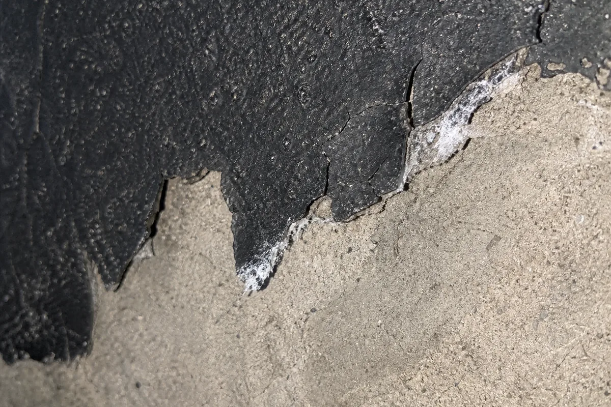 A close, well-lit view of an aging asphalt-emulsion foundation coating that has cracked, lifted, and flaked away from the concrete surface beneath it