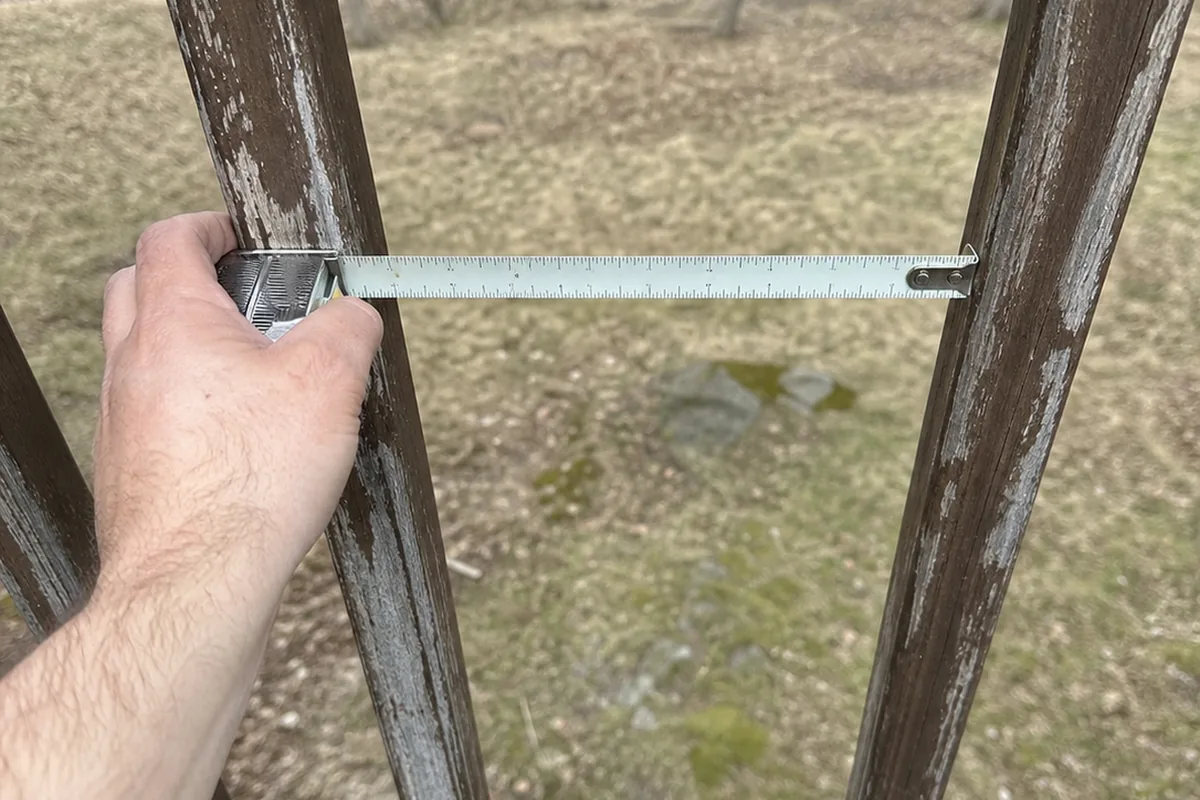Hand holding a metal tape measure stretched across the gap between two weathered deck balusters, the blade reading wider than four inches
