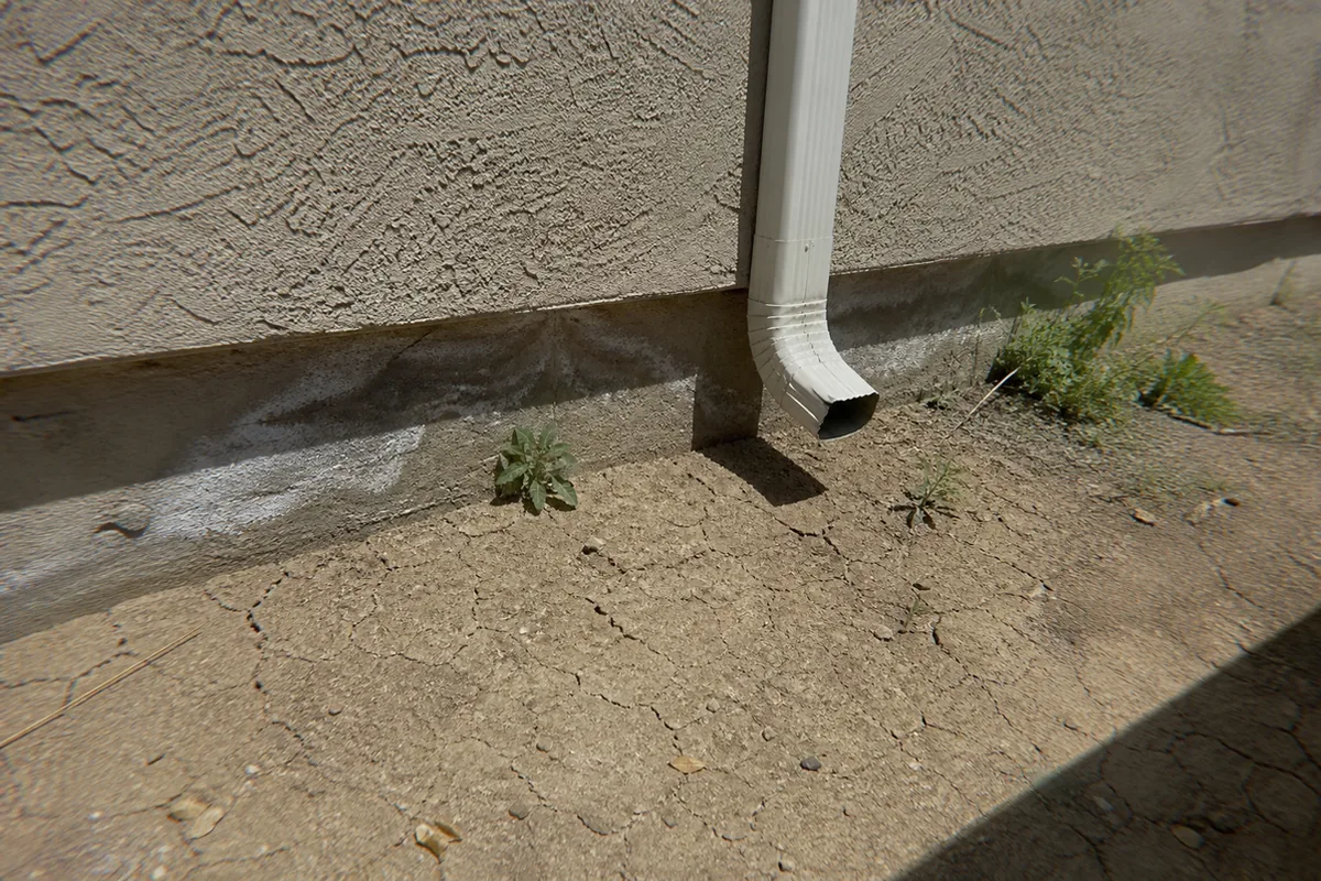 Downspout discharging directly onto sun-baked, cracked dirt against a stucco foundation