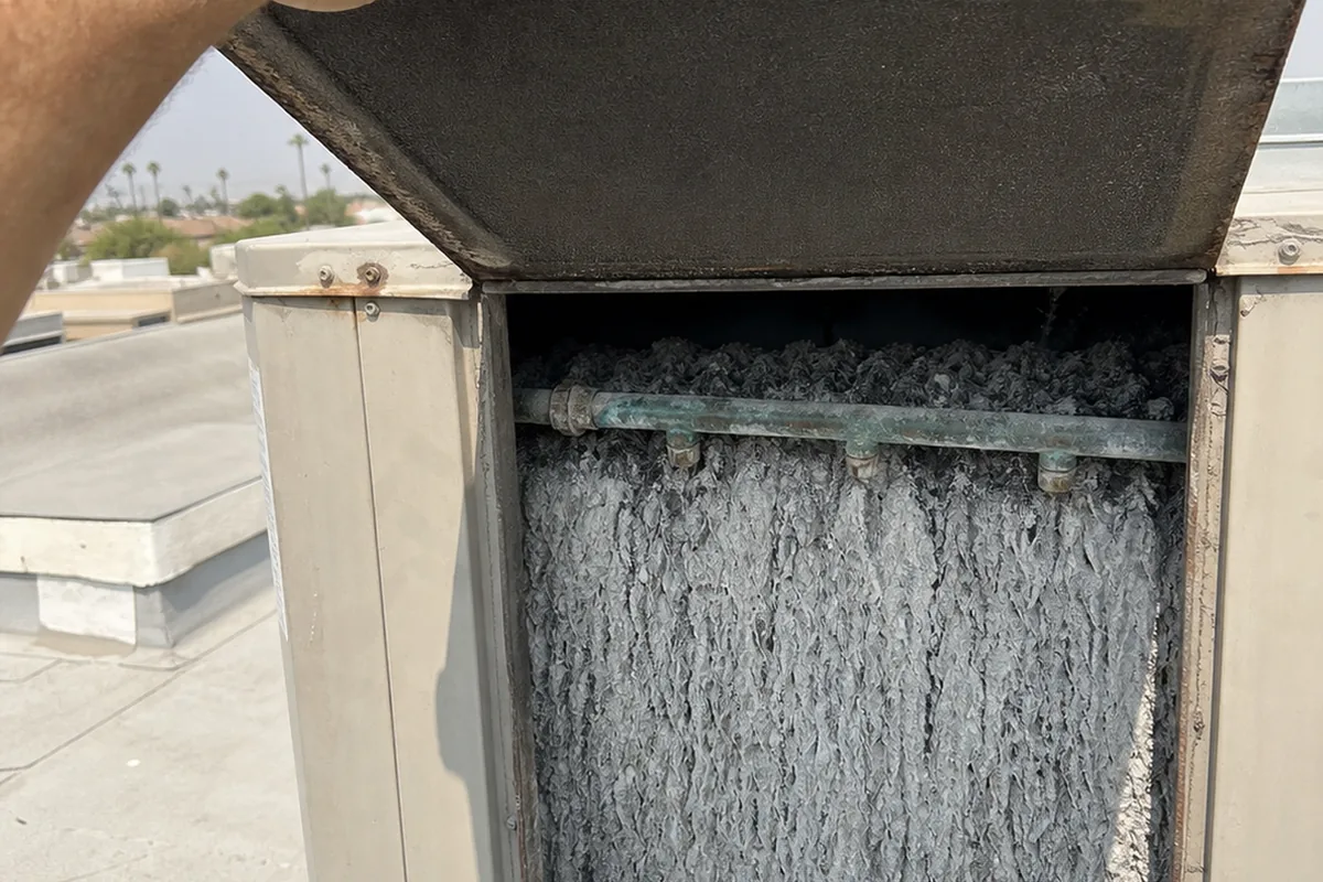 A technician's hand lifting the access panel on an evaporative cooler cabinet to reveal a mineral-scaled cooling pad and a corroded water distribution line inside
