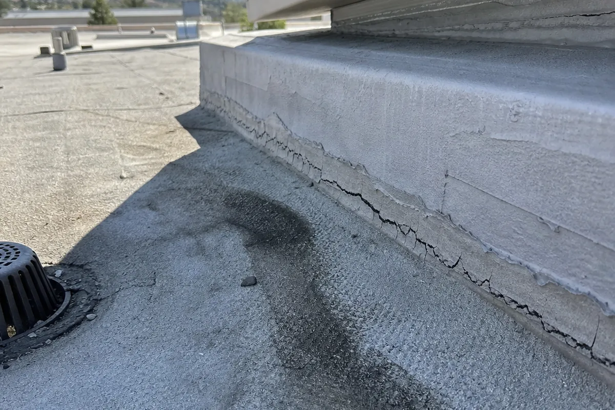 A low-angle view along a flat roof at the curb where a roof-mounted evaporative cooler meets its base, showing cracked flashing sealant and a dark stain trailing toward a roof drain