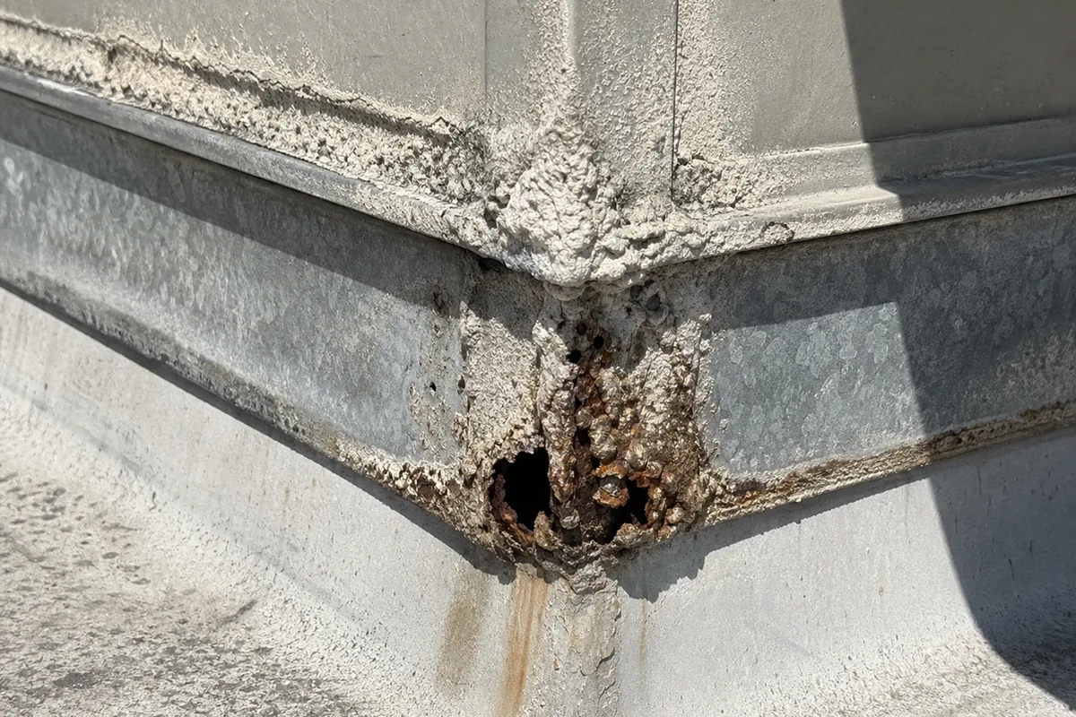 Extreme macro of a rooftop evaporative cooler cabinet's lower seam with thick mineral scale and rusted-through pinhole corrosion at the corner joint