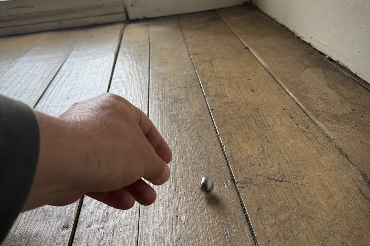 Home inspector's hand rolling a marble across an old wood floor toward a baseboard corner to check for a floor slope