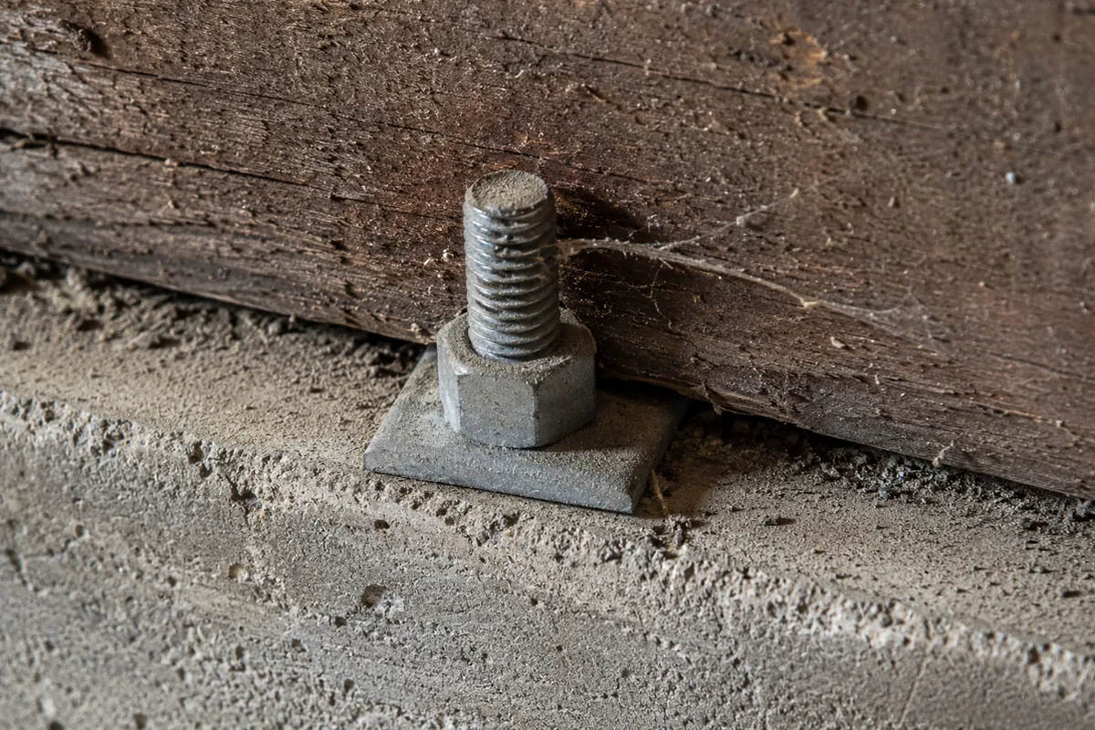 Close-up of a wood mudsill resting on a concrete foundation with a steel anchor bolt and washer, in a home crawlspace