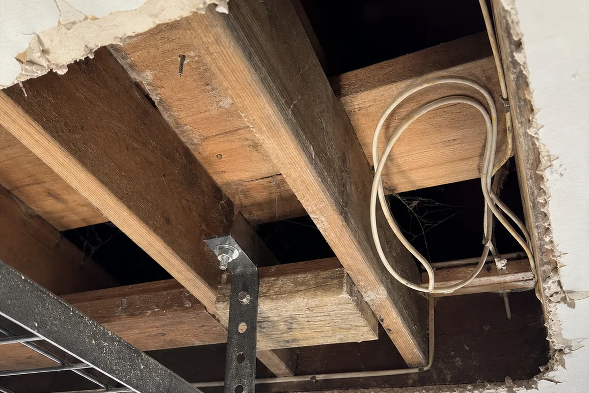 Straight-up view of a garage ceiling with a section of drywall cut away around a lag-bolted storage rack, exposing bare wood joists