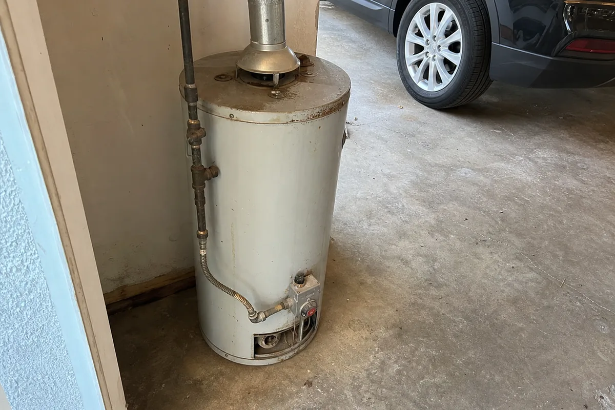 Older gas water heater sitting flat on a garage floor with no raised platform, a car parked a few feet away in the same bay