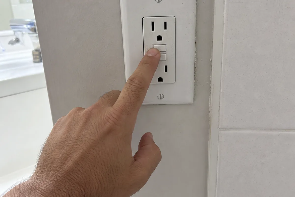 A bare finger pressing the test button on a white bathroom GFCI receptacle beside a tiled surround