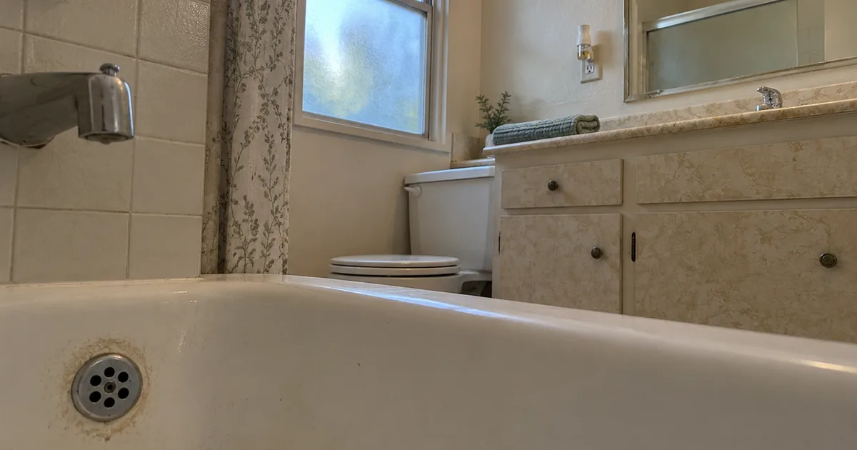 A seldom-used guest bathroom seen from tub height, with a dusty stained ring around the tub drain plate and a plug-in air freshener in the wall outlet above the vanity