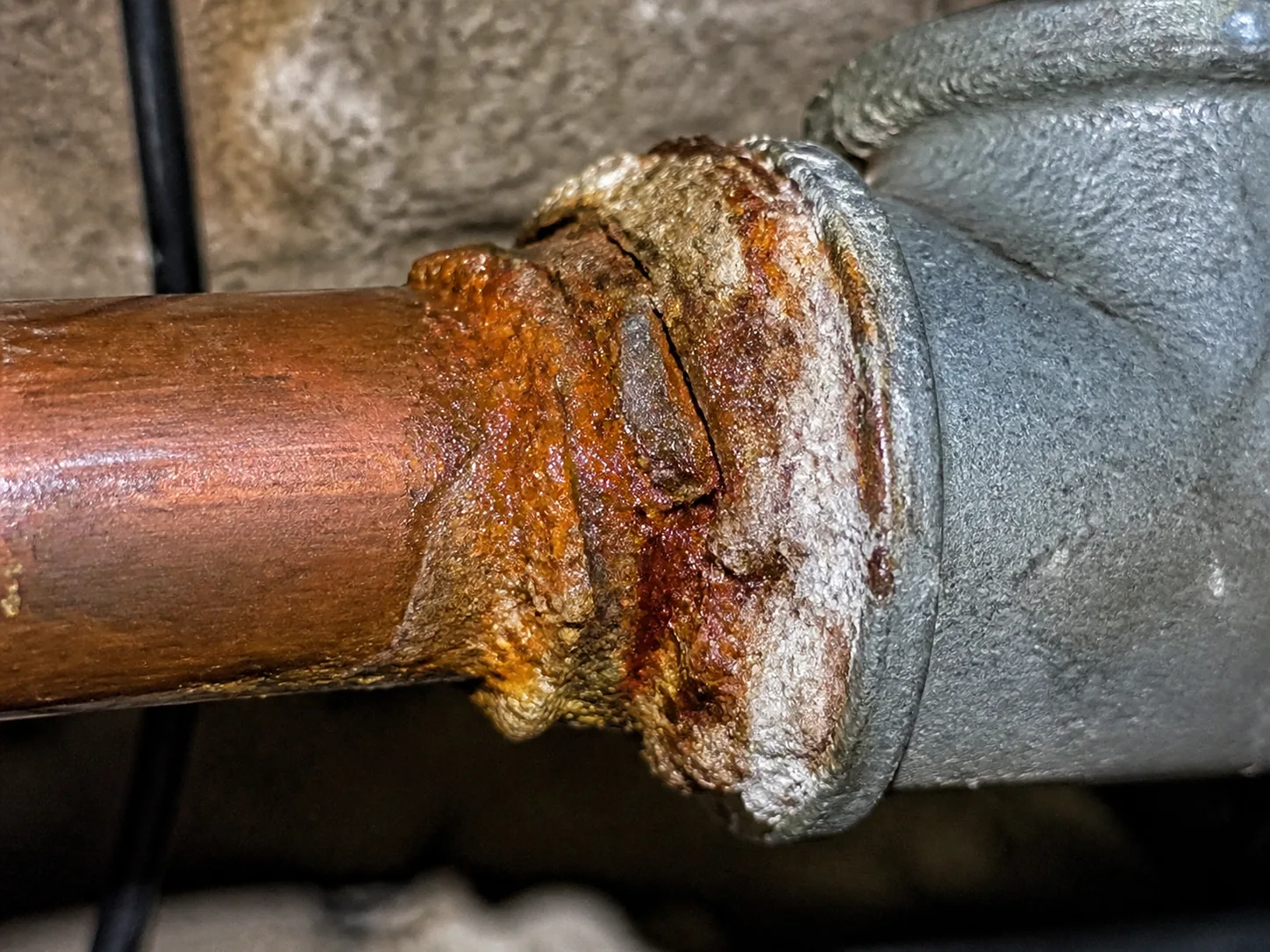 Extreme macro of a threaded transition where copper pipe meets galvanized steel, showing a thick crust of rust and white mineral bloom concentrated at the junction with clean pipe on either side