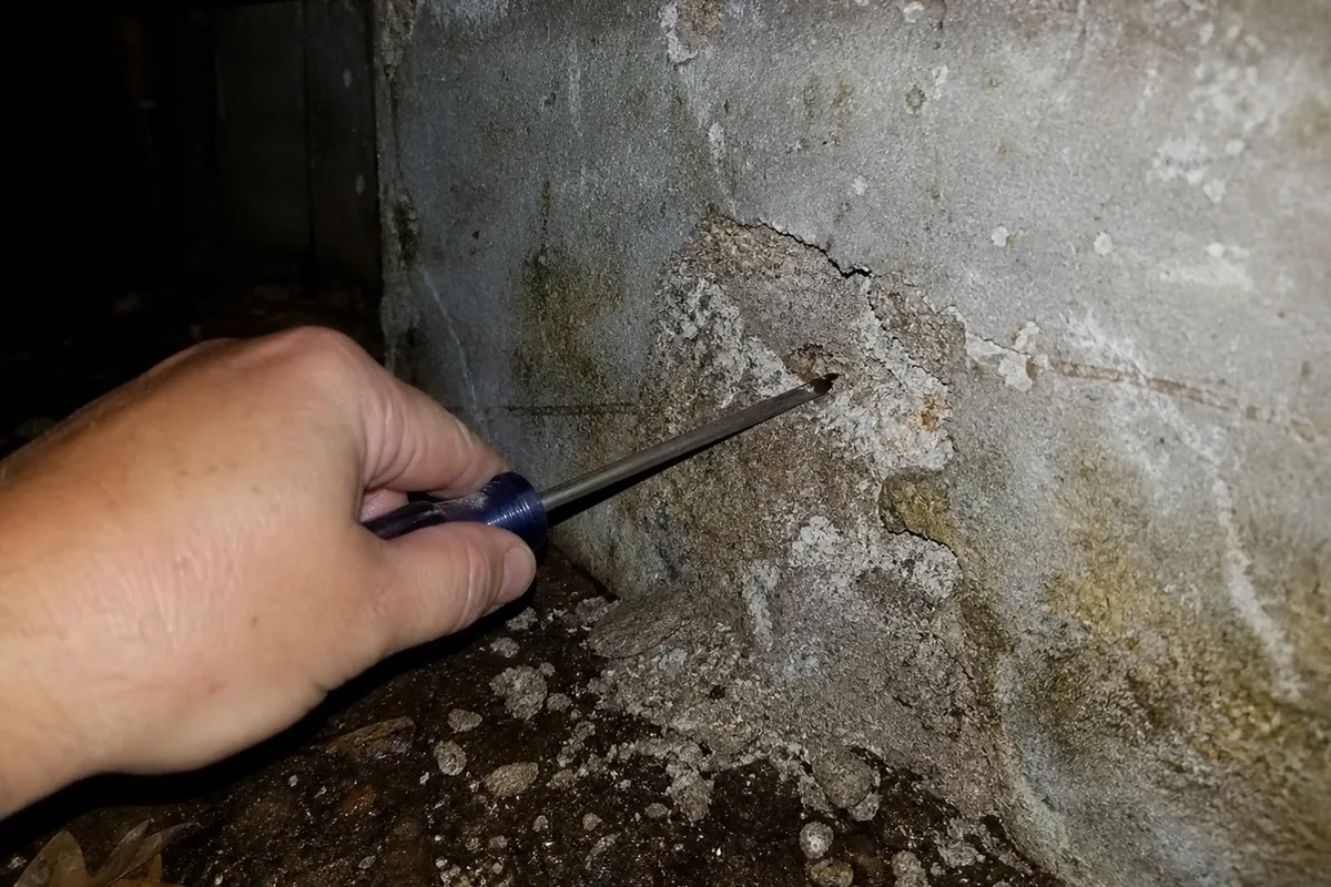 A hand pressing a screwdriver tip against a hollow-sounding spalled section of a foundation stem wall in a crawlspace, checking for soft or delaminated concrete