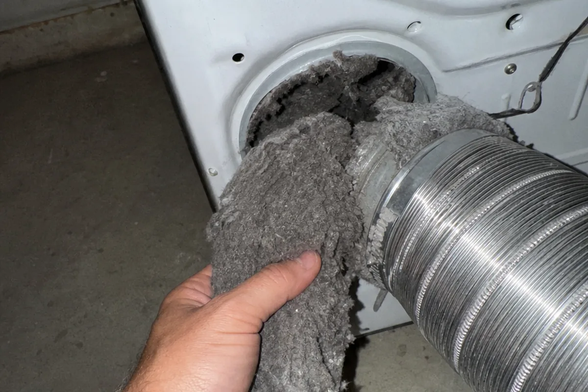 A thick mat of gray lint packed into the junction where a dryer transition duct meets the wall, the corrugated foil duct visibly deteriorated