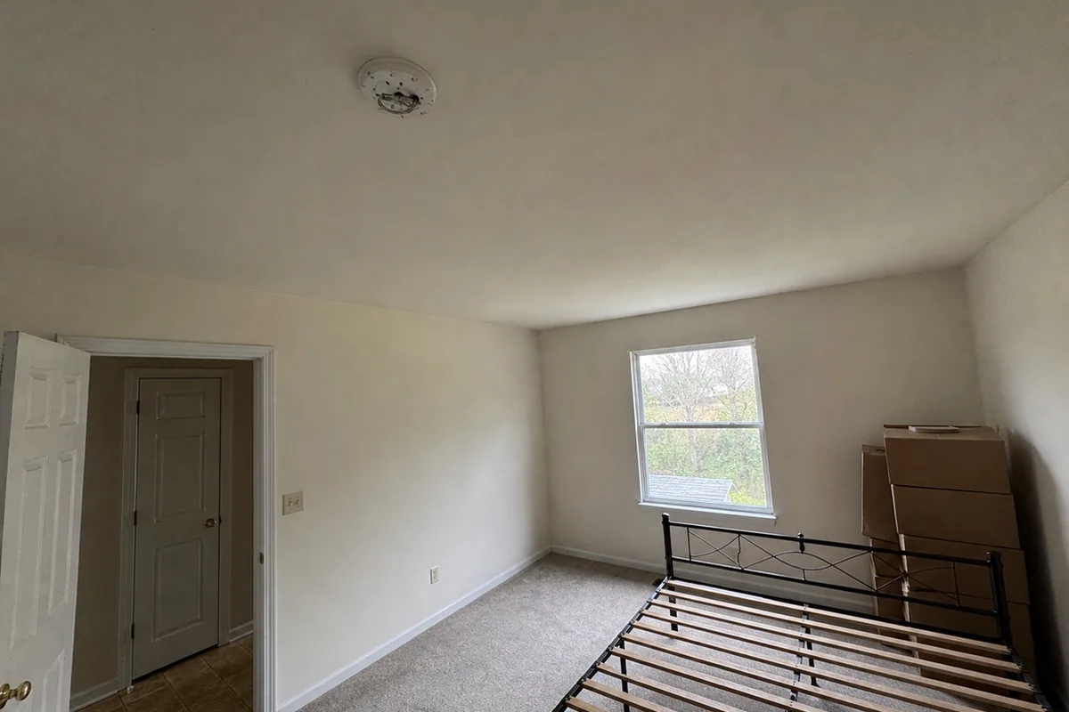 Wide interior view of a plainly finished converted bedroom, looking up at a bare ceiling with an empty round alarm mounting bracket and a short curl of wire, no alarm attached, flat overcast light through an uncurtained window