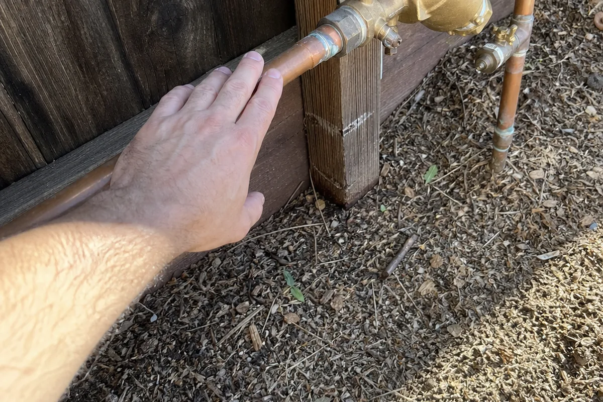 A hand tracing an exterior copper irrigation supply line to document its connection to a backflow prevention assembly at a Moreno Valley property