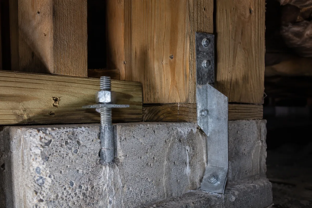 Crawlspace close-up of a galvanized anchor bolt through a sill plate beside a steel seismic hold-down bracket