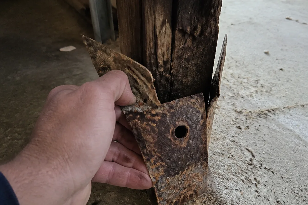 A bare hand pulling a badly rust-corroded metal post base bracket away from the bottom of a wood post, exposing stained wood behind the connector