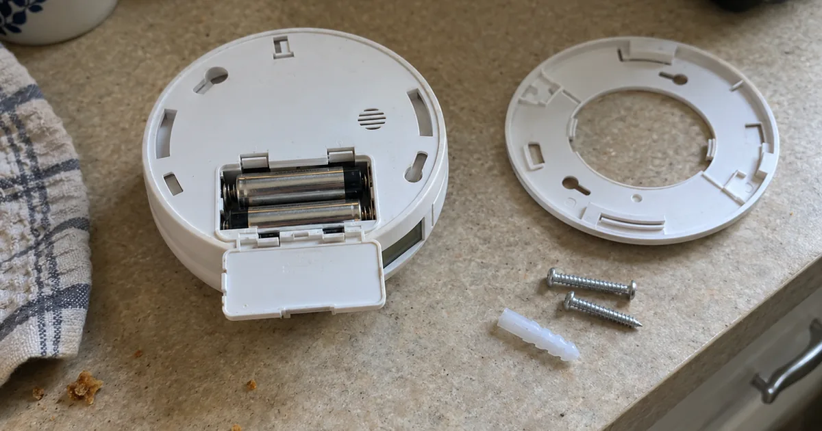 A white combination smoke and carbon monoxide detector lying face down on a kitchen counter with its battery door open and two AA batteries inside, the round mounting plate, two screws and a wall anchor beside it
