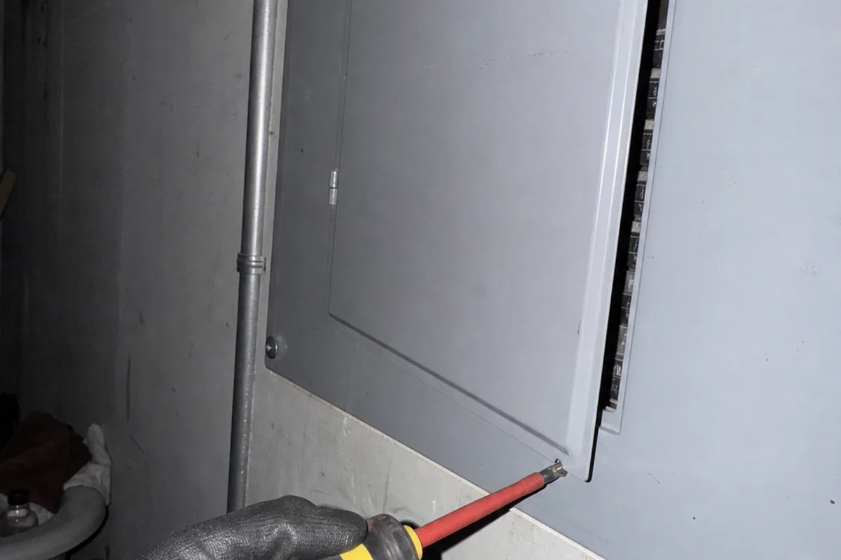 A wide shot of an opened panel during inspection with a gloved hand holding a voltage tester near the breaker row, panel cover leaning against the wall