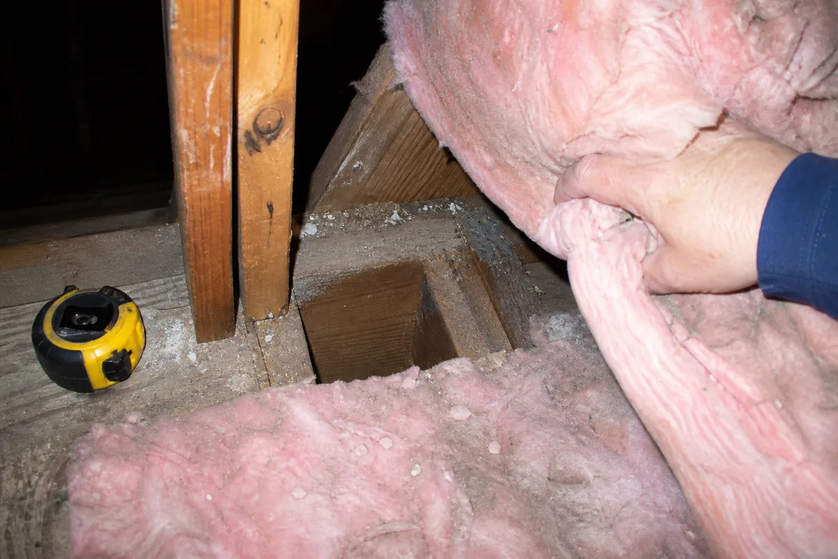 A hand pulling back attic insulation to expose an open, unblocked stud bay at the top plate near an addition's tie-in point, tape measure set down in frame