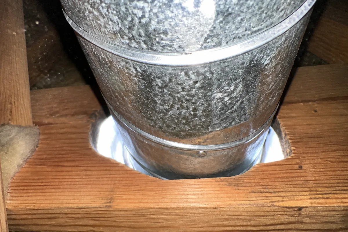 Macro view of a large unsealed gap around an HVAC duct penetrating a top plate in a Paramount attic, daylight visible around the duct into the room below