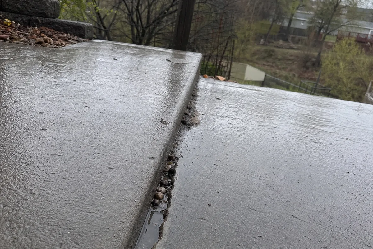 Low ground-level view of a concrete patio slab in a back yard with a wet, open separation crack running roughly parallel to a slope edge, the two sides sitting at slightly different heights, a leaning wooden fence post visible beyond in gray overcast light