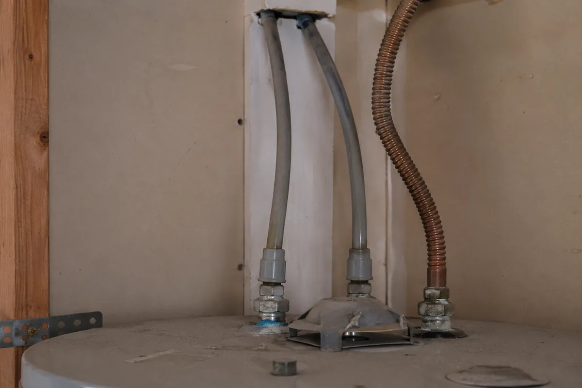 Gray polybutylene supply lines running from the water heater up into the wall cavity of an early-1990s home, the typical install pattern an inspector traces