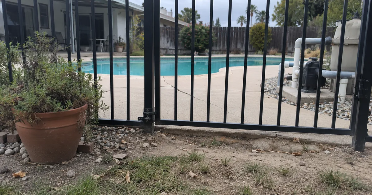 A black tubular steel pool gate seen from outside at ground level, with the latch mounted low on the post, eroded dirt leaving a gap under the gate, a terracotta planter beside the fence, and the pool, a cracked concrete deck and the pump pad behind it