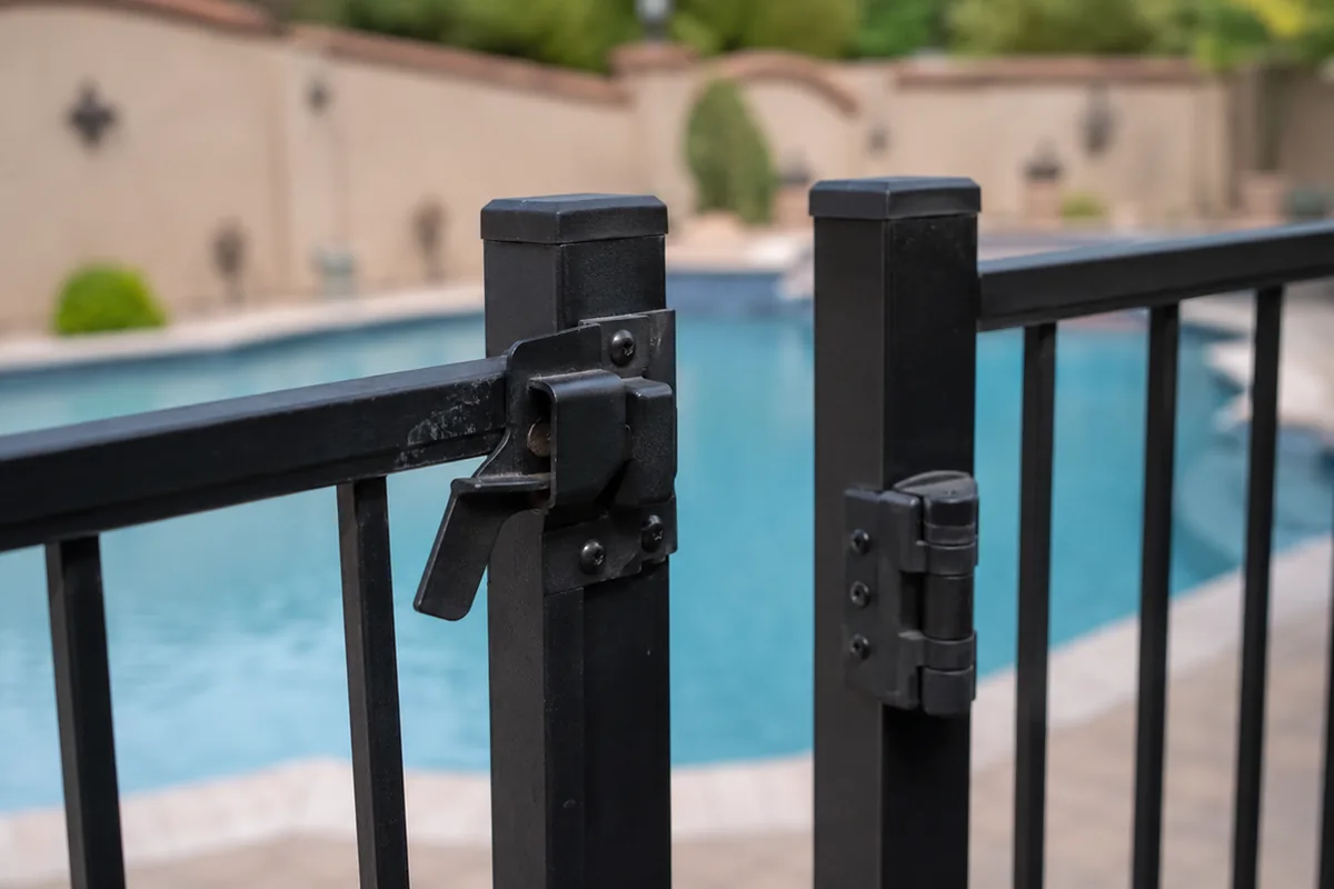 Close-up of a residential pool safety gate latch and self-closing hinge on a metal fence, the barrier hardware a buyer must confirm works