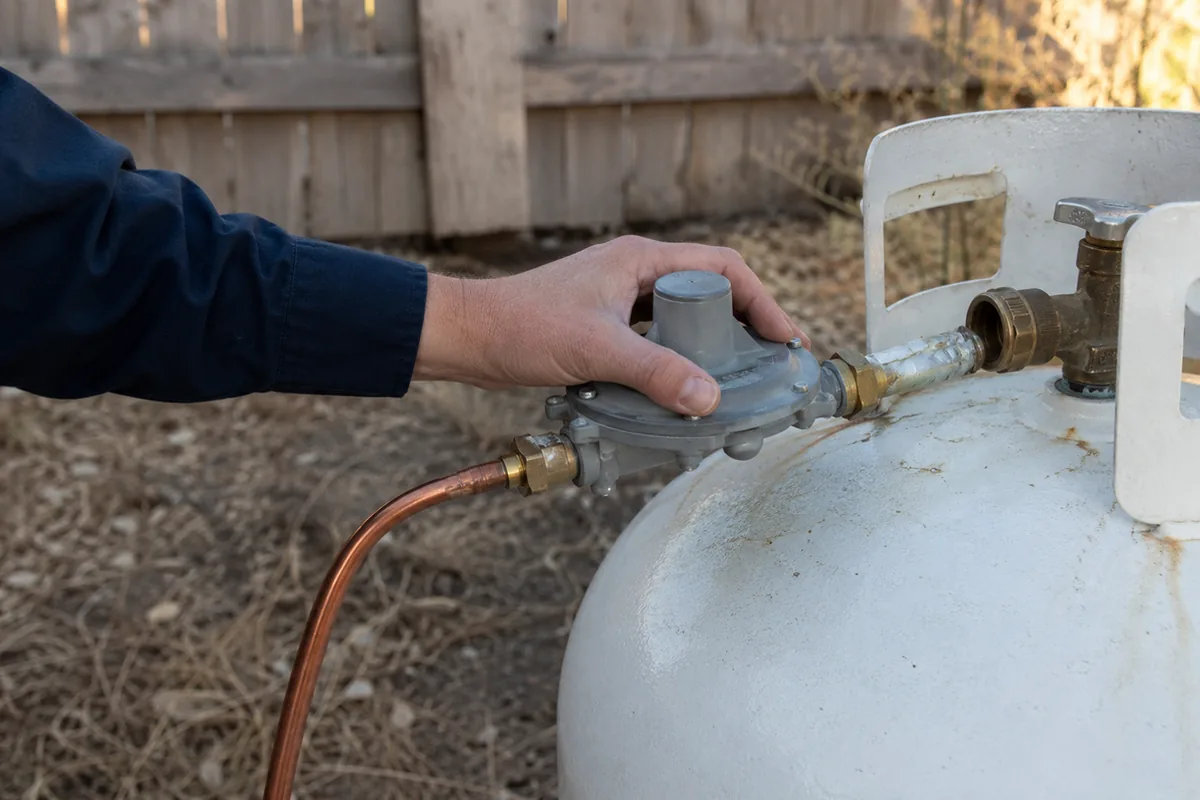 Home inspector examining an above-ground propane tank regulator and supply line at the side yard of a rural home