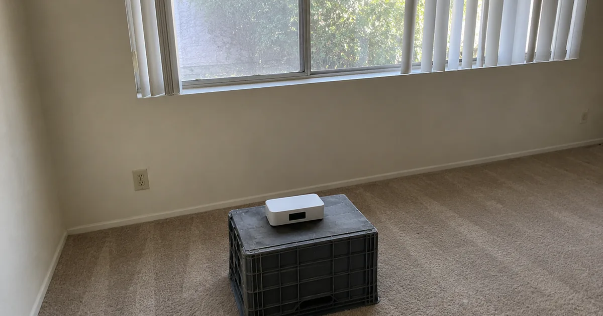 A small white continuous radon monitor set on an upturned crate about 20 inches off the carpet in an empty ground-floor room, away from the closed slider window and walls