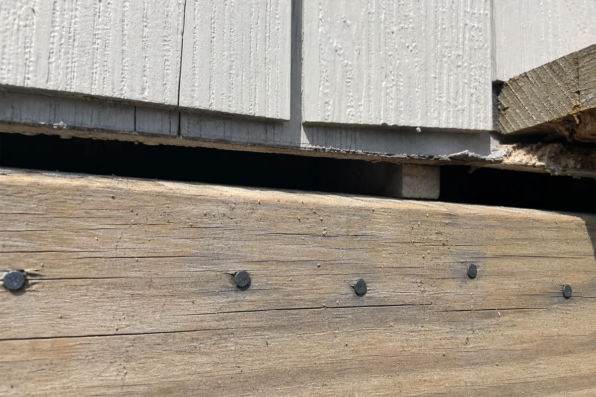 Close-up of a ledger board joint showing a visible daylight gap between the ledger and the house framing, with plain nail heads instead of bolts