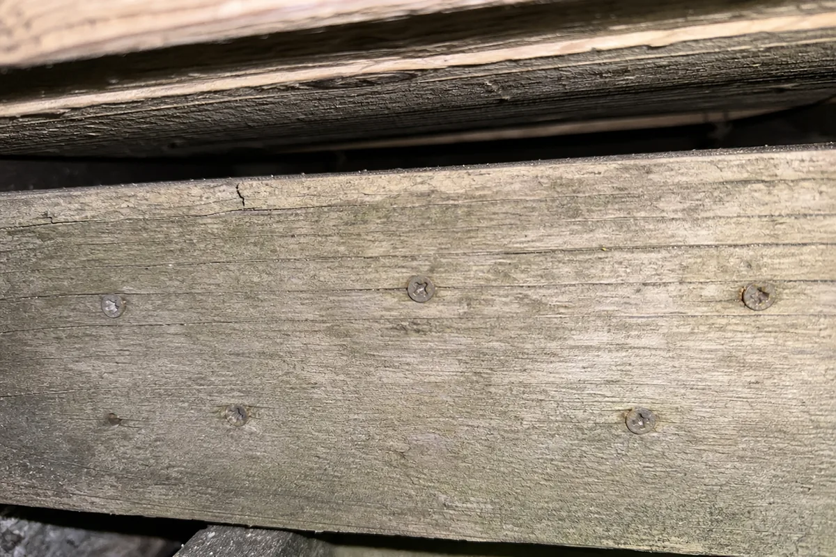 Close macro of a weathered deck ledger board fastened with small deck-screw heads instead of structural bolt heads, with a narrow gap at the top edge where flashing should be