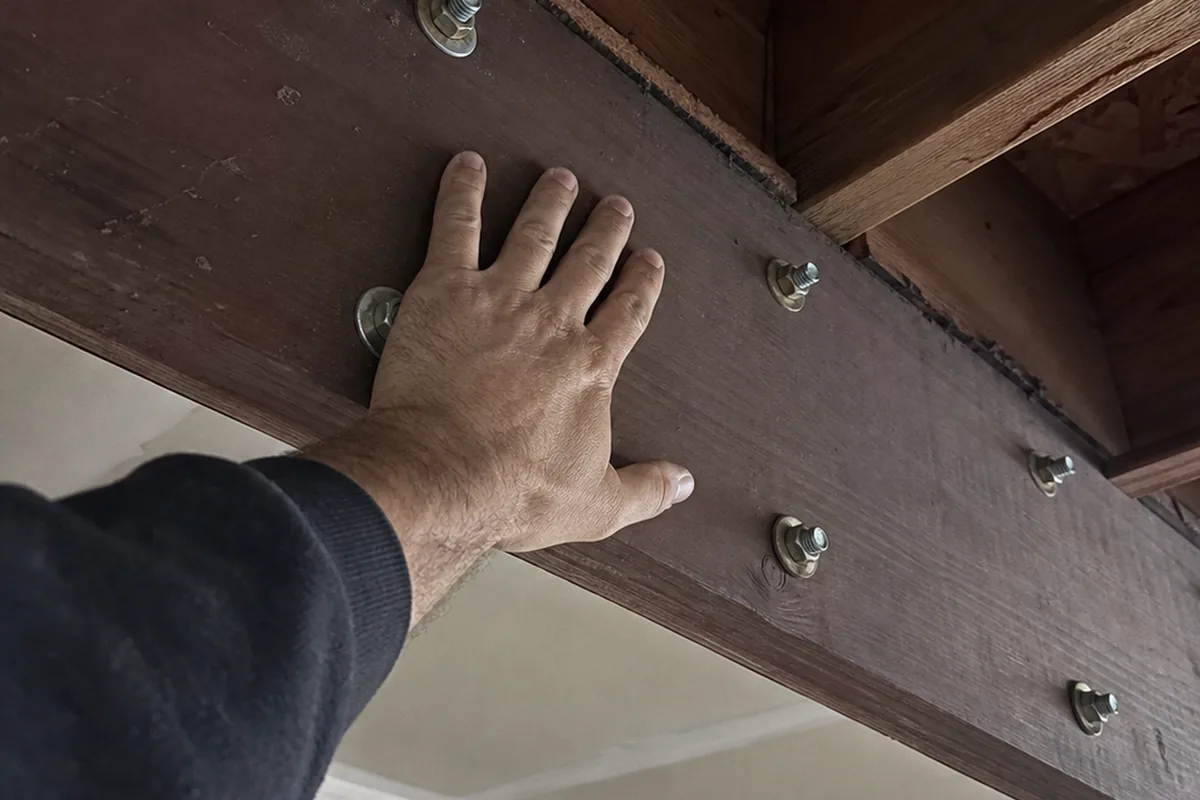 A hand pressing against the underside of a deck ledger board near a row of through-bolts, checking the connection for movement from below