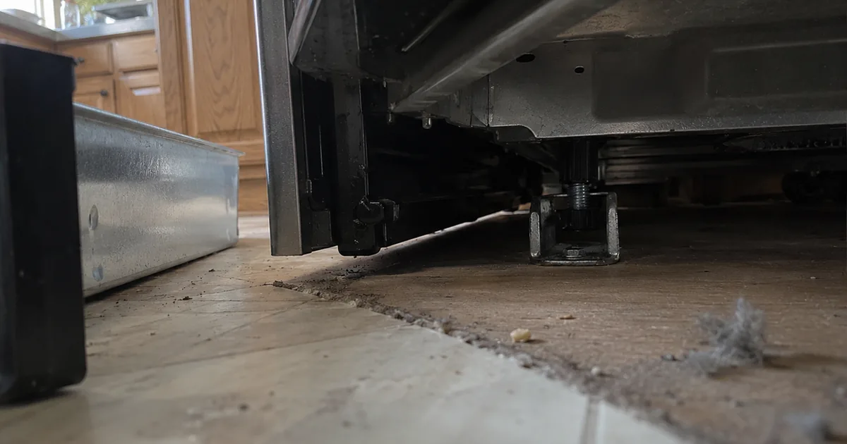 Floor-level view under a freestanding kitchen range with its drawer pulled out, a rear leg seated in a steel anti-tip bracket