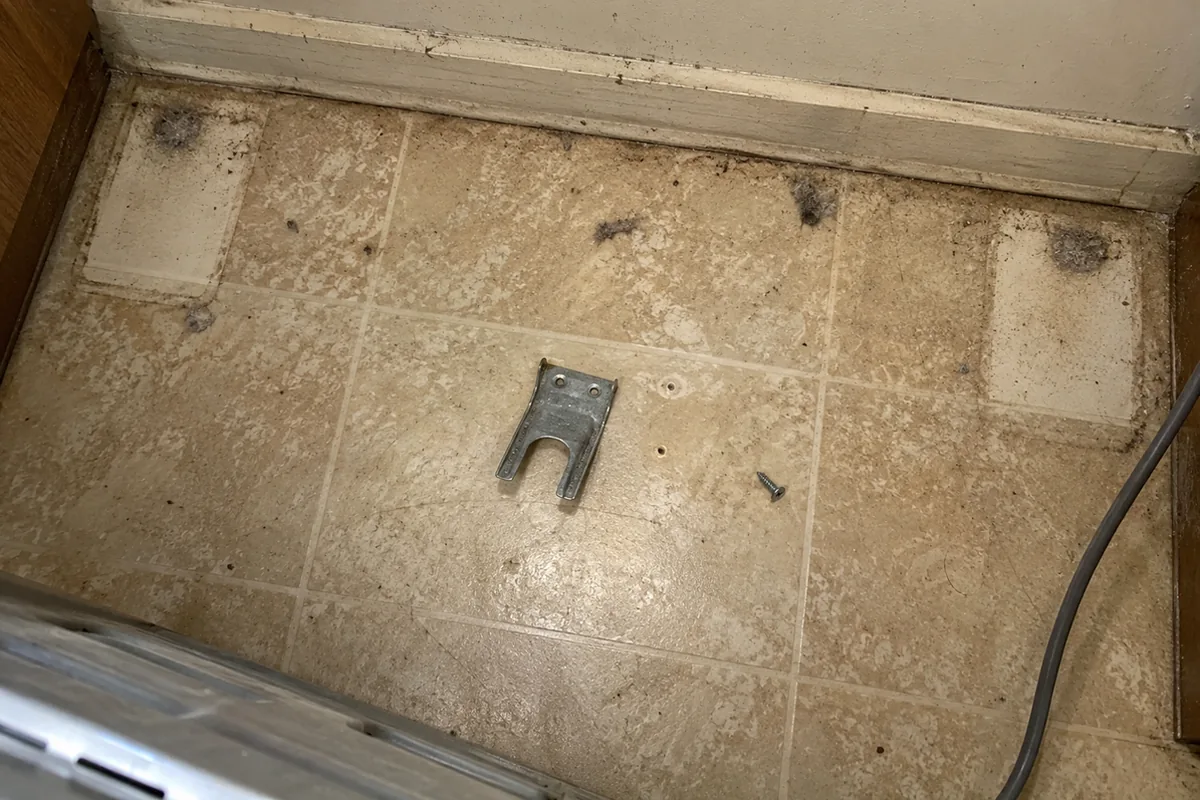 A loose fork-shaped anti-tip bracket and screw lying on a kitchen floor behind a pulled-out range, with empty screw holes beside it