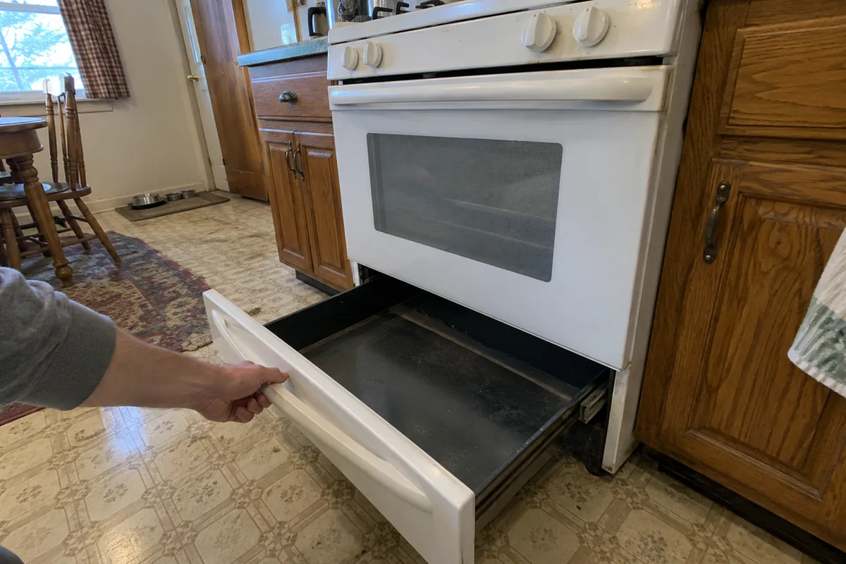 A hand pulling the bottom storage drawer fully out of a white freestanding gas range in a dated kitchen