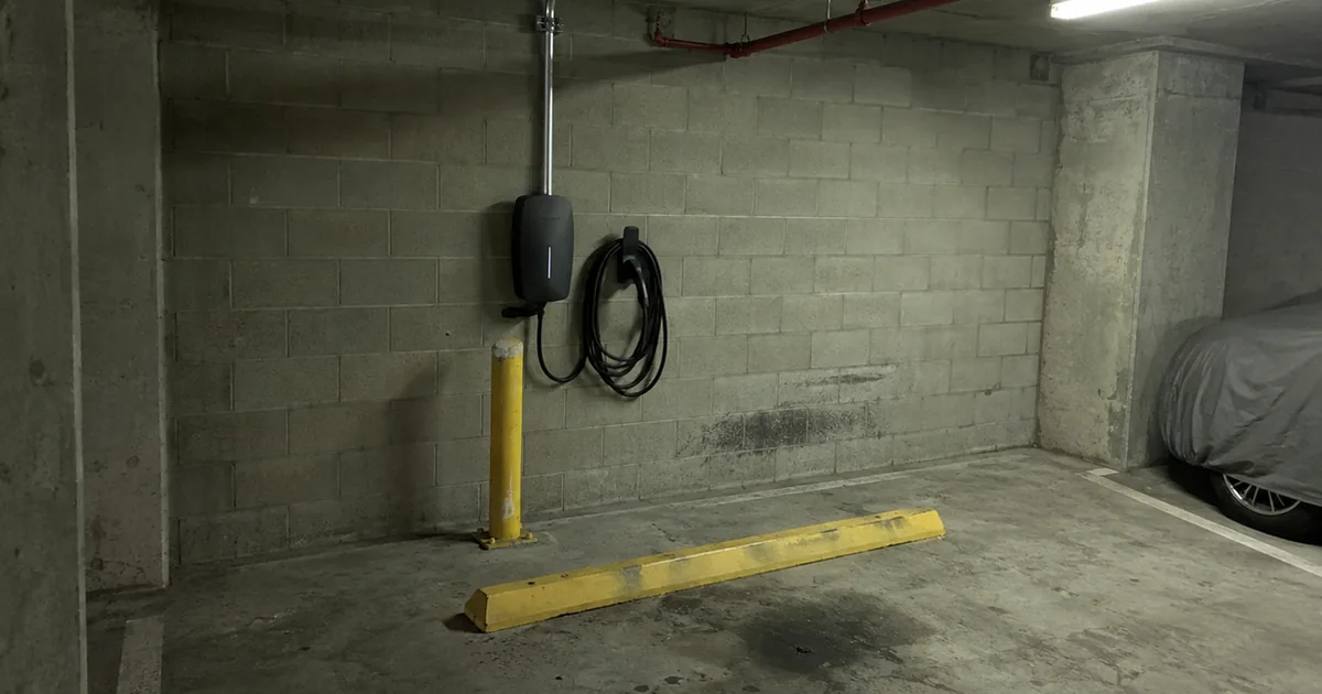 Wall-mounted EV charger at an empty assigned stall in a condo parking garage, with metal conduit running up the block wall to the ceiling, a yellow bollard, a wheel stop, and an oil stain on the slab
