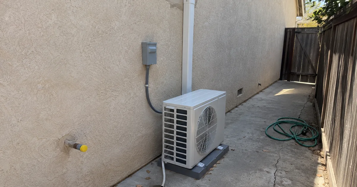 New heat pump outdoor unit on a plastic pad in a narrow stucco side yard, with a wall disconnect, a white line-set cover, a loose condensate tube, and a capped gas pipe stub where the old gas service was