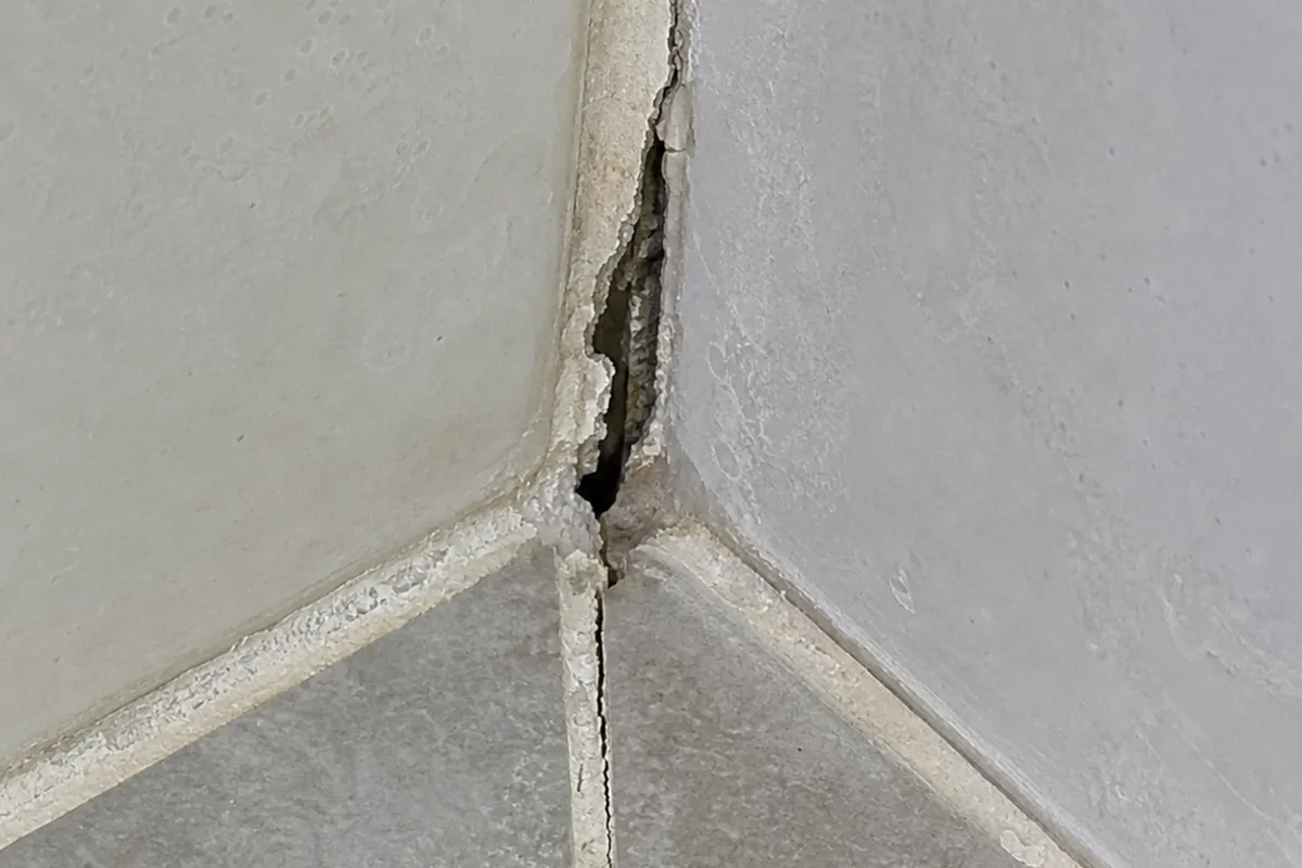 Cracked and missing grout in the corner of a tile shower where the floor meets the wall, the gap open to the mortar bed behind it
