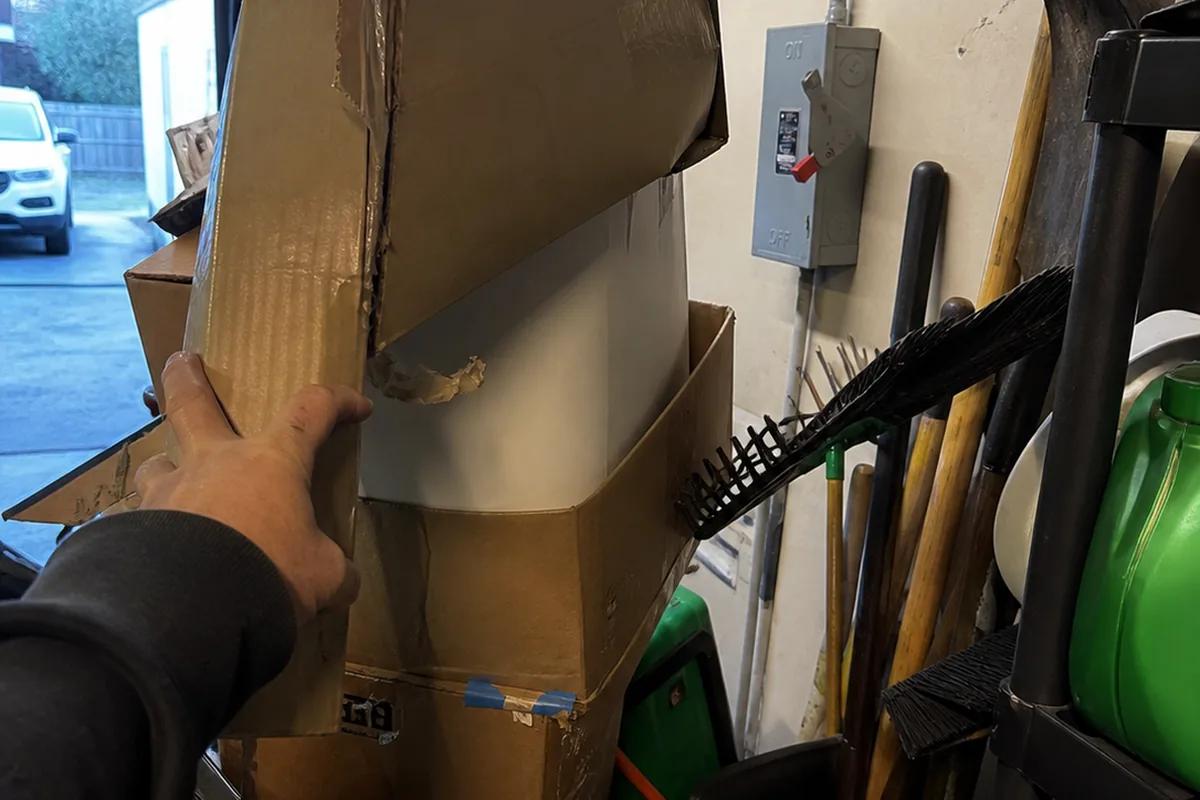 A hand pulling back stored boxes and yard tools from in front of a garage-mounted electrical disconnect to check clearance, dim mixed garage light