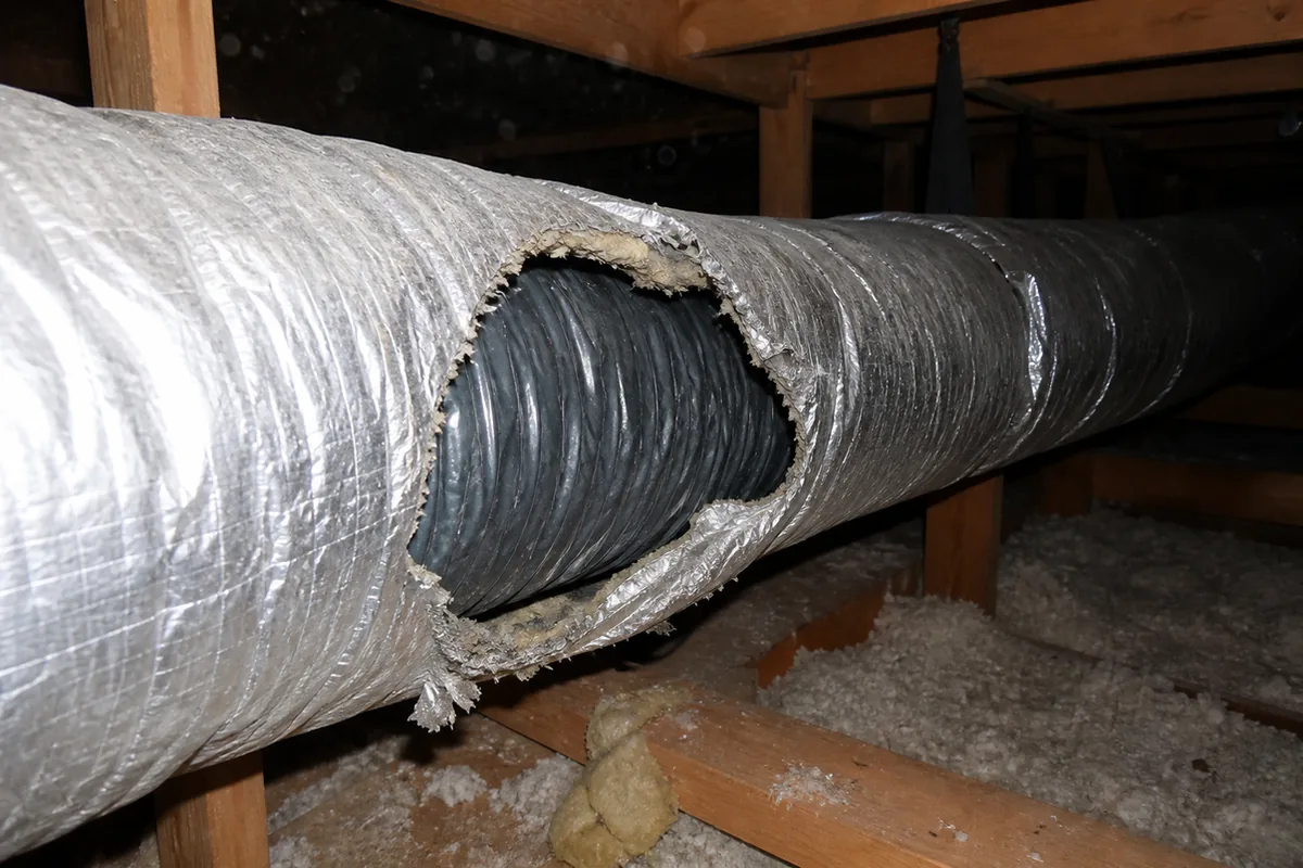 Wide attic view of a supply duct run with torn, gapped insulation wrap under harsh flash, dusty and dim