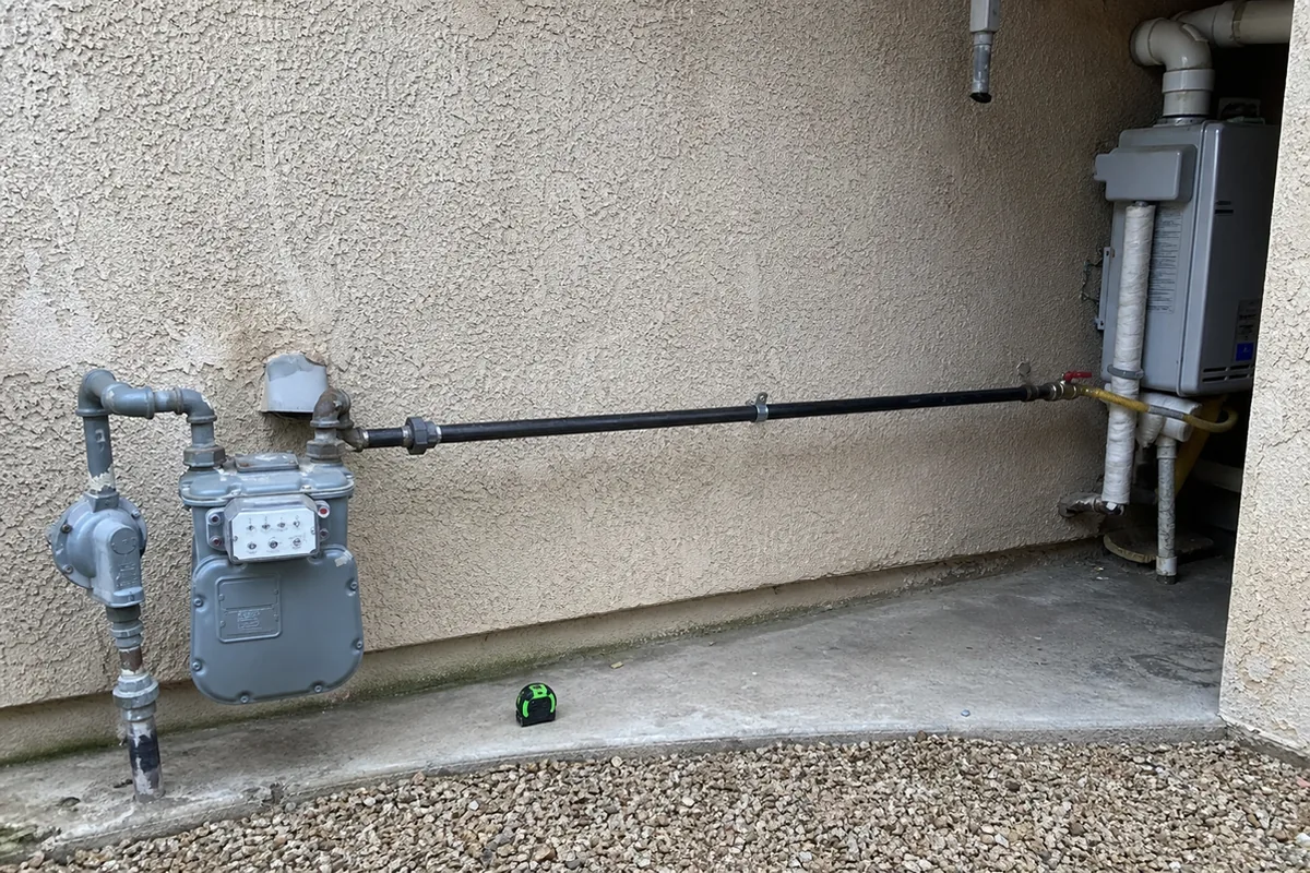 A black iron gas line running along an exterior stucco wall from a gas meter to a tankless water heater installation, with a tape measure resting on the ground nearby